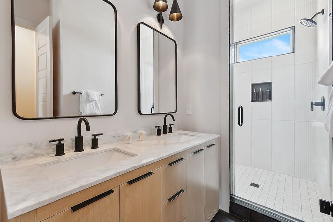 Sleek modern bathroom with double-sink marble countertop, light wood vanity and matte black faucets, two rounded-rectangle mirrors, and a glass-enclosed walk-in shower with white tile and a narrow horizontal window.