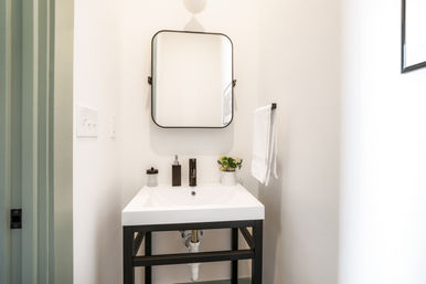 Bright minimalist powder room with a white rectangular sink on a black metal vanity, rounded-corner mirror, wall towel, soap dispenser and small potted plant.
