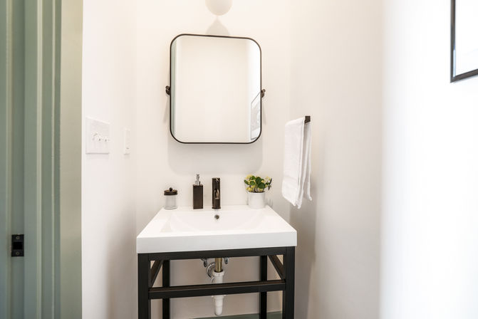 Bright minimalist powder room with a white rectangular sink on a black metal vanity, rounded-corner mirror, wall towel, soap dispenser and small potted plant.
