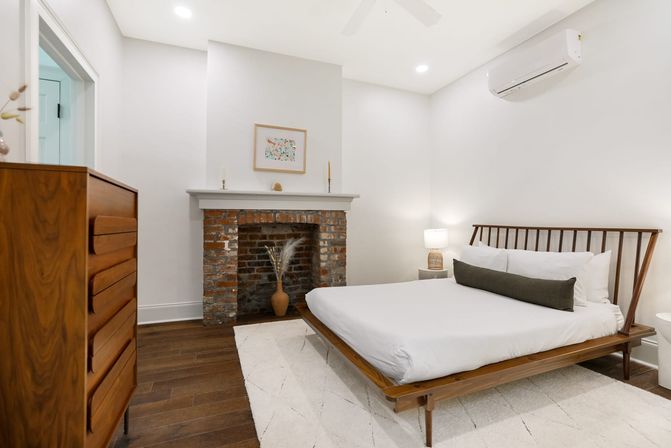 Minimalist mid-century modern bedroom with low wooden platform bed, white linens and dark bolster, exposed brick fireplace with decorative vase, walnut dresser, hardwood floors, neutral rug and wall-mounted air conditioner.