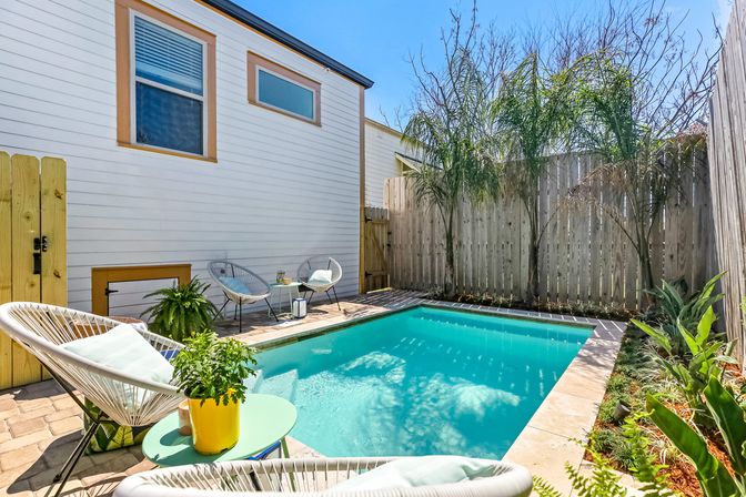 Sunny backyard plunge pool with turquoise water beside a white modern house, wood privacy fence, palm trees, potted plants and wicker lounge chairs on a cozy patio