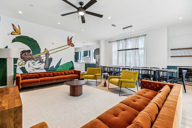 Bright modern apartment clubhouse lounge with rust-orange velvet sofas and mustard-yellow armchairs around a round wooden coffee table, large mural of a trombone player with tropical leaves and butterflies on a white wall, floor-to-ceiling windows, long communal dining table, open kitchen and ceiling fan in a light-filled open-concept space.