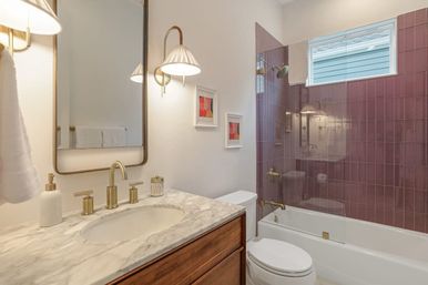 Bright residential bathroom with marble countertop and wooden vanity, brass fixtures, glass-enclosed tub/shower with mauve subway tiles and small high window.
