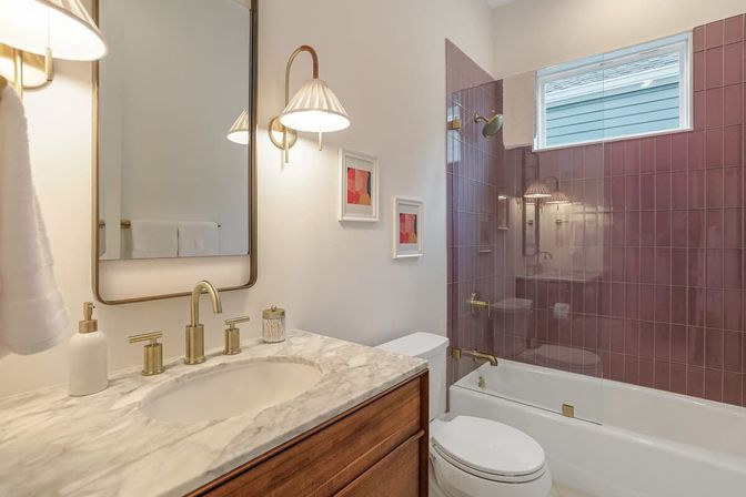 Bright residential bathroom with marble countertop and wooden vanity, brass fixtures, glass-enclosed tub/shower with mauve subway tiles and small high window.