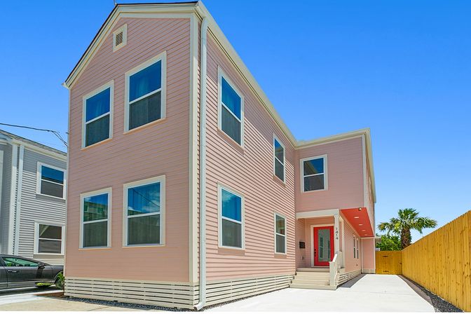 Bright pink two-story coastal-style house with modern siding, red front door, concrete driveway and a palm tree under a clear blue sky.