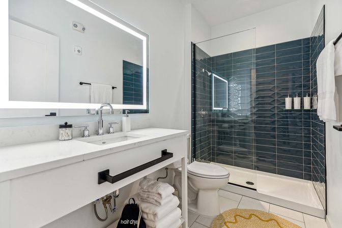 Sleek modern bathroom with backlit LED mirror, white quartz vanity and chrome fixtures, glass walk-in shower with navy subway tile, stacked towels and round yellow bath mat