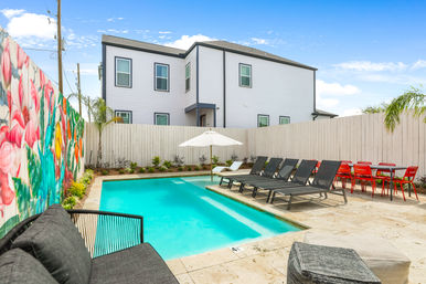Modern urban townhouse backyard with a rectangular turquoise swimming pool, colorful floral mural on the left fence, black lounge chairs and white umbrella on the deck, red outdoor dining chairs, small palm plants and sunny blue sky.