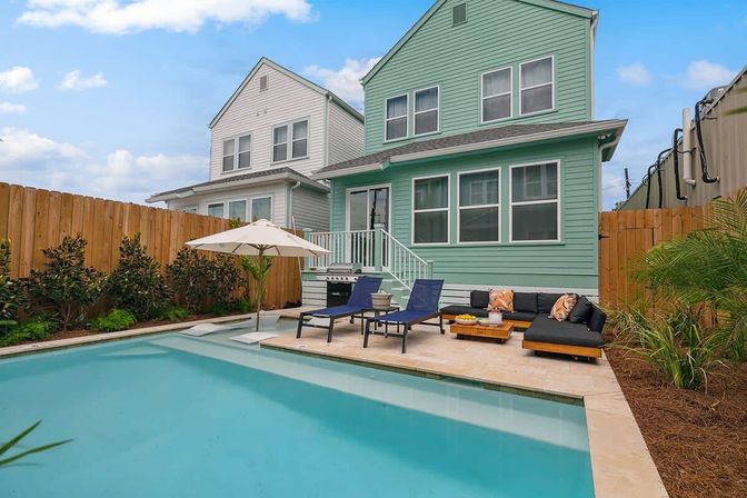 Cheerful turquoise two-story home with a rectangular backyard pool, sun loungers, umbrella and cozy outdoor sofa beside a wooden fence.