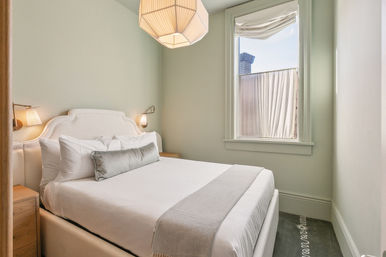 Cozy small urban guest bedroom with soft green walls, white upholstered headboard, crisp white linens, gray bolster and throw, pendant light and wall sconces, and a window with a partial city view.