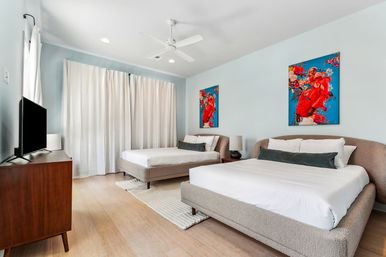 Bright, breezy modern vacation rental bedroom with two upholstered platform beds, light-blue walls, colorful red artwork, ceiling fan, flat-screen TV on a wooden dresser, sheer white curtains and hardwood floors.