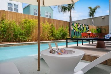 Sunny backyard pool scene with a shallow turquoise plunge, white lounge chair and umbrella, modern ice bowl holding chilled bottles, palm trees and bamboo along a wooden fence, and a colorful mural with an orange outdoor dining set in the background.