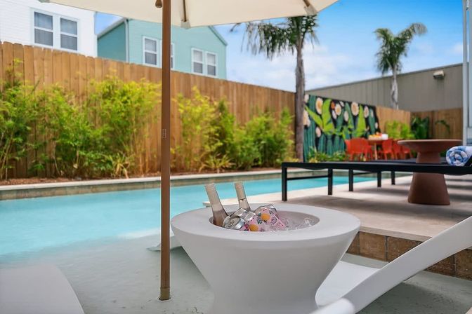 Sunny backyard pool scene with a shallow turquoise plunge, white lounge chair and umbrella, modern ice bowl holding chilled bottles, palm trees and bamboo along a wooden fence, and a colorful mural with an orange outdoor dining set in the background.