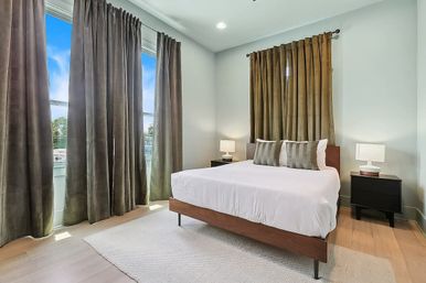Sunlit modern bedroom in an urban apartment: wooden platform bed with white linens and striped accent pillows, matching black nightstands and lamps, tall green floor-length curtains over large windows, light hardwood floors and neutral area rug.
