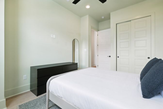 Bright minimalist bedroom with white bed and navy pillows, black low dresser and arched mirror against soft green walls, ceiling fan and double closet doors.