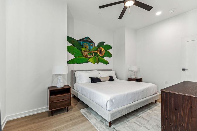 Bright modern bedroom interior with a low gray platform bed dressed in white linens, mid-century wooden nightstands and lamps, a colorful tropical banana-leaf wall mural featuring a small house and brass instrument, ceiling fan, hardwood floors and dresser.