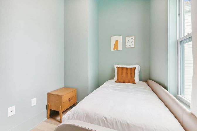 Cozy compact modern bedroom with mint-green walls, single white bed, rust-orange accent pillow, wooden nightstand and bright window light.