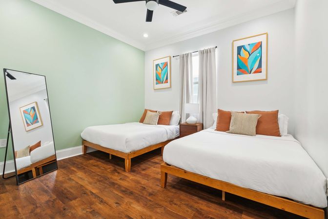 Cozy modern guest room with two twin beds on wooden frames, white bedding, warm hardwood floors, mint accent wall, colorful botanical prints, full-length mirror, ceiling fan, and neutral curtains.