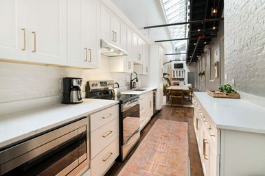 Bright modern white galley kitchen with brass hardware and stainless-steel appliances, skylight with exposed beams, dark wood floors and a pink runner leading to a dining area.