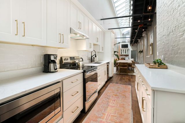 Modern white galley kitchen in an urban loft with a skylight and black exposed beams, brass cabinet pulls, stainless steel appliances, marble-look countertops, dark hardwood floors, a pink patterned runner, and an open dining area at the far end.