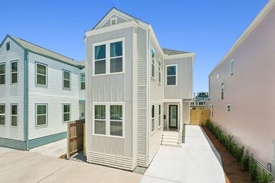 Modern two-story townhouse exterior with beige vertical siding and white trim, narrow concrete driveway, small fenced yard and neighboring homes under a clear blue sky