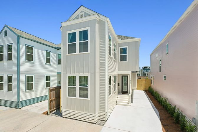 Modern two-story townhouse exterior with beige vertical siding and white trim, narrow concrete driveway, small fenced yard and neighboring homes under a clear blue sky