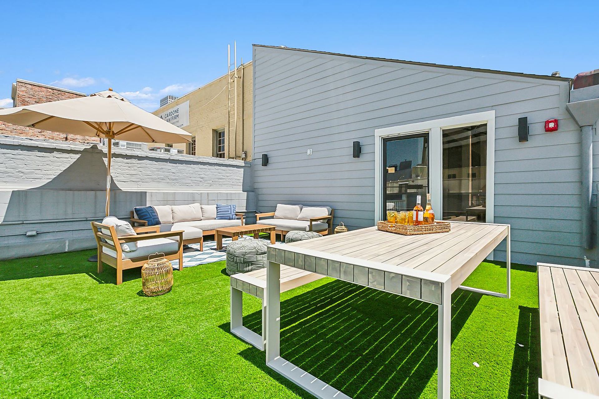 Sunlit urban rooftop patio with bright artificial turf, wooden dining table and benches, cushioned lounge seating under a large beige umbrella, striped poufs and wicker accents against a gray-sided building and clear blue sky.