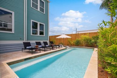 Private backyard lap pool beside a teal coastal-style two-story house with four black lounge chairs, wooden side tables, a white patio umbrella, bamboo and palm plants, and a wooden privacy fence under a blue sky.