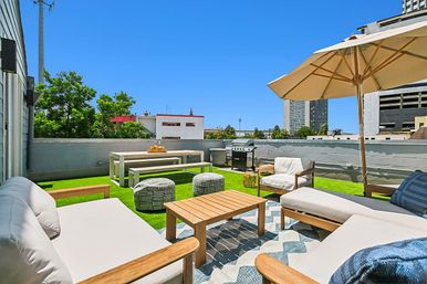 Sunny urban rooftop terrace with cushioned wooden lounge chairs, umbrella, coffee table, picnic bench, grill and green turf against a blue sky and city skyline.