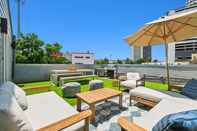 Sunny urban rooftop terrace with cushioned wooden lounge chairs, umbrella, coffee table, picnic bench, grill and green turf against a blue sky and city skyline.