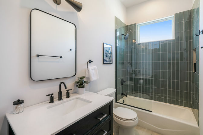 Contemporary residential bathroom with white vanity, black fixtures and framed mirror, potted plant, toilet, and glass-enclosed tub-shower with vertical green tiles and a high window