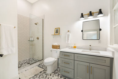 Bright modern residential bathroom with glass-enclosed subway-tile shower, gray vanity with brass hardware and white countertop, wall mirror and triple sconce, wicker-topped toilet, and patterned hexagon tile floor — stylish bathroom remodel design.