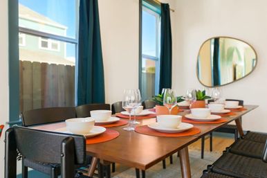 Sunlit modern dining room with a wooden table set for guests — white bowls and plates on orange placemats, wine glasses, small potted succulents, black chairs, teal curtains, and an oval wall mirror.