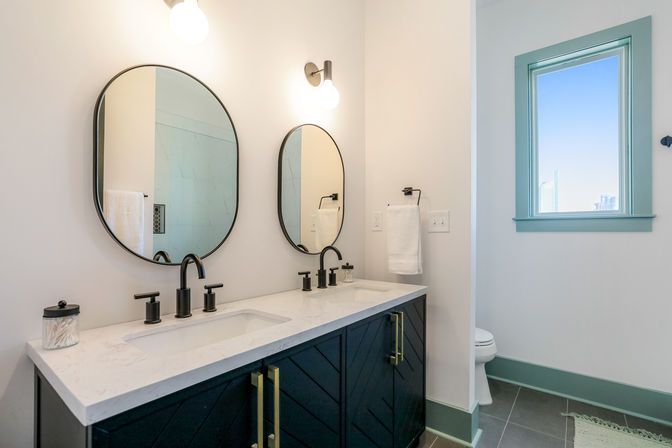 Modern coastal-inspired bathroom with white quartz double-sink vanity, matte-black faucets, oval mirrors, dark blue cabinets with brass pulls, teal-trimmed window and gray tile floor.