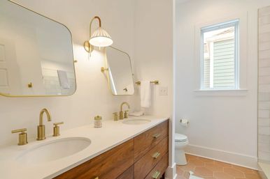 Bright modern bathroom with double sink white countertop, brass faucets and mirrors, walnut wood vanity, hex terracotta floor tiles and a sunlit window.