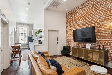 Stylish urban loft living room with exposed red brick accent wall, wall-mounted TV, tan leather sofa with navy pillows, glass coffee table, green accent chair, media console, open white kitchen with bar stools, hardwood floors and a large window overlooking city buildings