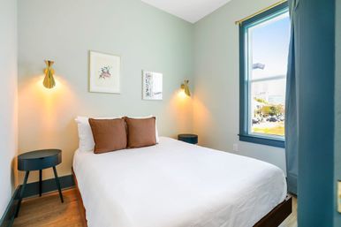 Cozy modern bedroom with white bedding and two brown pillows, sage-green walls, brass wall sconces, teal window trim, round dark bedside tables, wood floors and a sunny city skyline view through the window