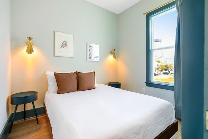 Cozy modern bedroom with white bedding and two brown pillows, sage-green walls, brass wall sconces, teal window trim, round dark bedside tables, wood floors and a sunny city skyline view through the window