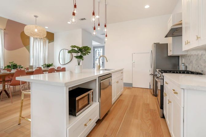 Bright open-concept modern kitchen with white island and quartz countertop, stainless steel appliances, light wood floors, hanging pendant bulbs, potted plant and adjacent dining area with blush chairs and abstract wall mural.