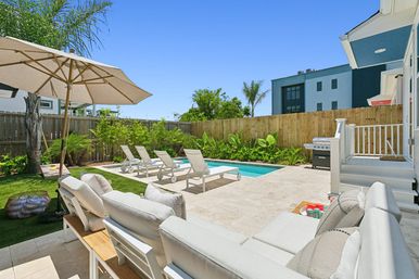 Sunny modern backyard with a rectangular swimming pool, four white lounge chairs, cushioned patio sofa under a large umbrella, stone tile terrace, barbecue grill, and tropical landscaping behind a wooden fence.