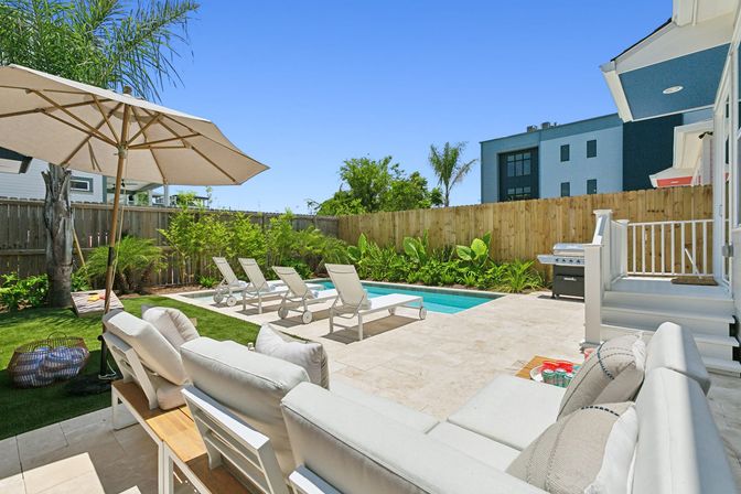 Sunny modern backyard with a rectangular swimming pool, four white lounge chairs, cushioned patio sofa under a large umbrella, stone tile terrace, barbecue grill, and tropical landscaping behind a wooden fence.