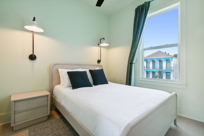Bright minimalist bedroom with white queen bed, navy pillows, wall-mounted lamps, light wood nightstand and a window view of a blue two-story building with decorative balconies