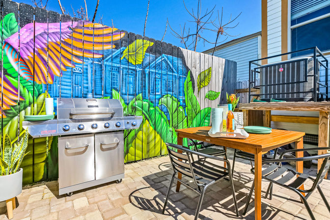 Sunny urban backyard patio with stainless steel gas grill, wooden dining table and metal chairs on paver stones, vibrant tropical mural painted on wooden fence