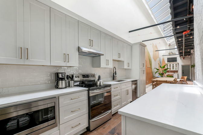 Bright modern white open-plan loft kitchen with stainless steel appliances, quartz countertops, subway-tile backsplash, large skylight and exposed black ceiling beams opening to a dining area with plants.