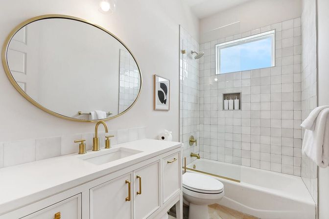 Bright modern white bathroom with gold hardware, oval mirror above a white vanity with undermount sink, and tiled tub-shower with glass panel and high window