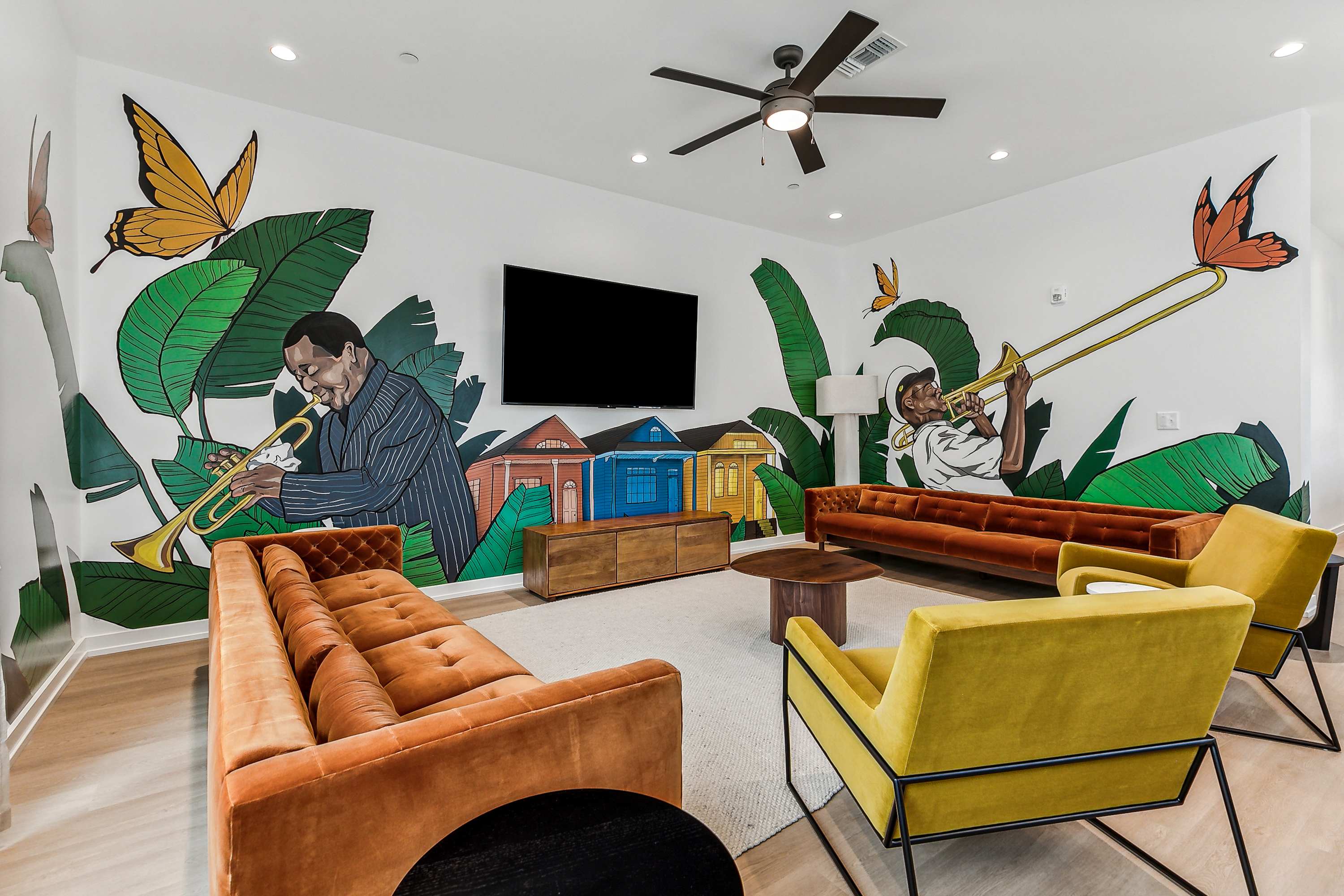 Lively lounge with a large wall mural of illustrated jazz musicians playing trumpet and trombone among oversized tropical leaves, butterflies and colorful row houses; mid-century velvet orange sofas, yellow armchairs, wall-mounted TV and ceiling fan.