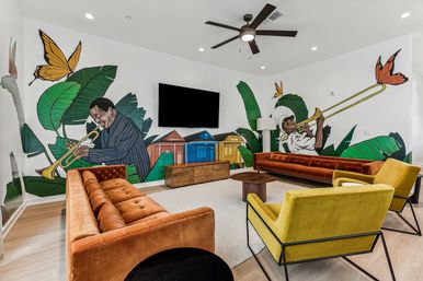 Lively lounge with a large wall mural of illustrated jazz musicians playing trumpet and trombone among oversized tropical leaves, butterflies and colorful row houses; mid-century velvet orange sofas, yellow armchairs, wall-mounted TV and ceiling fan.