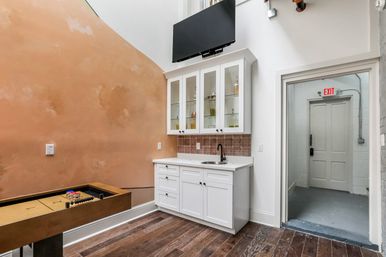 Stylish recreational room with white glass-front kitchenette and mini sink, wall-mounted TV above, peach textured accent wall, hardwood floors and a shuffleboard table beside a doorway with an exit sign.