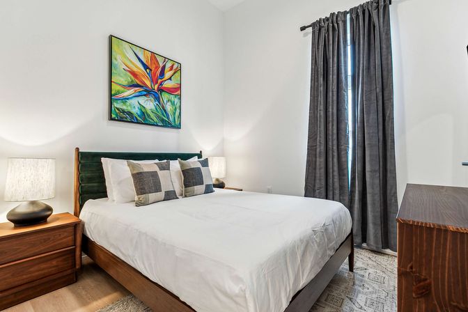 Modern bedroom interior with white walls, wooden bed frame and green velvet headboard, crisp white bedding with checkered throw pillows, wooden nightstand and lamp, colorful bird-of-paradise painting above the bed, tall gray curtains and a wooden dresser.