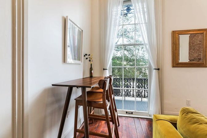 Cozy apartment nook with mid-century wooden bar table and stools, sheer-curtained tall window opening to ornate iron balcony, hardwood floors and a yellow sofa