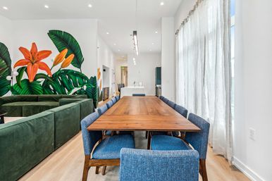 Bright modern open-plan dining room with long walnut table, blue upholstered chairs, green velvet sectional, large tropical wall mural featuring orange lilies, and sheer floor-to-ceiling curtains